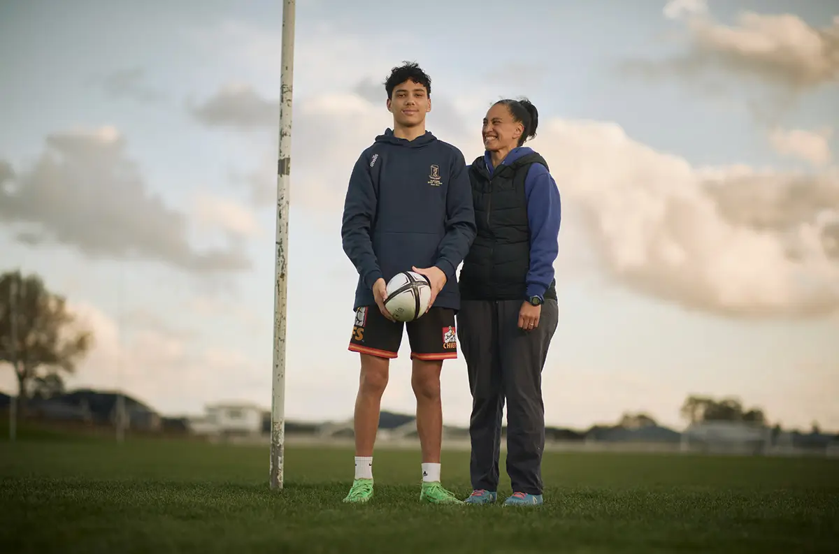 New Zealand bus driver Hikitia stands with her teenage son Cody on a rugby field. Cody holds a rugby ball while his mum smiles proudly.