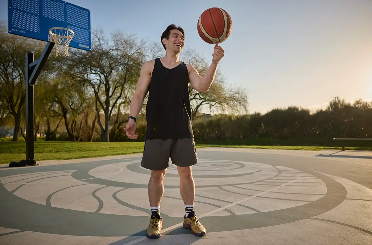Joshua, a Tauranga bus driver, smiling while spinning a basketball on his finger at an outdoor court.